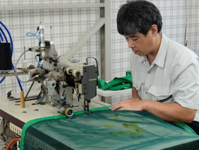 作業台で工業用ミシンを操作し、緑色のネットを縫い合わせる男性