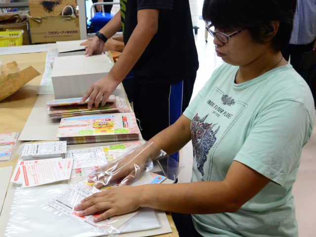 紙製品を透明な袋に詰めている女性とその周囲の作業台の風景