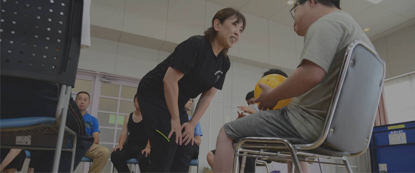 女性が椅子に座る男性に笑顔で話しかけ、黄色いボールを持つ男性が応じている様子