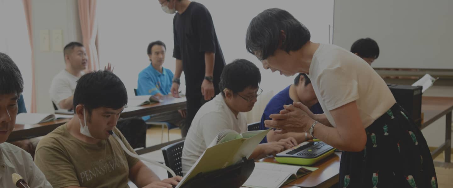 白いシャツの女性が机に向かう男性たちと話しながら活動をサポートしている様子