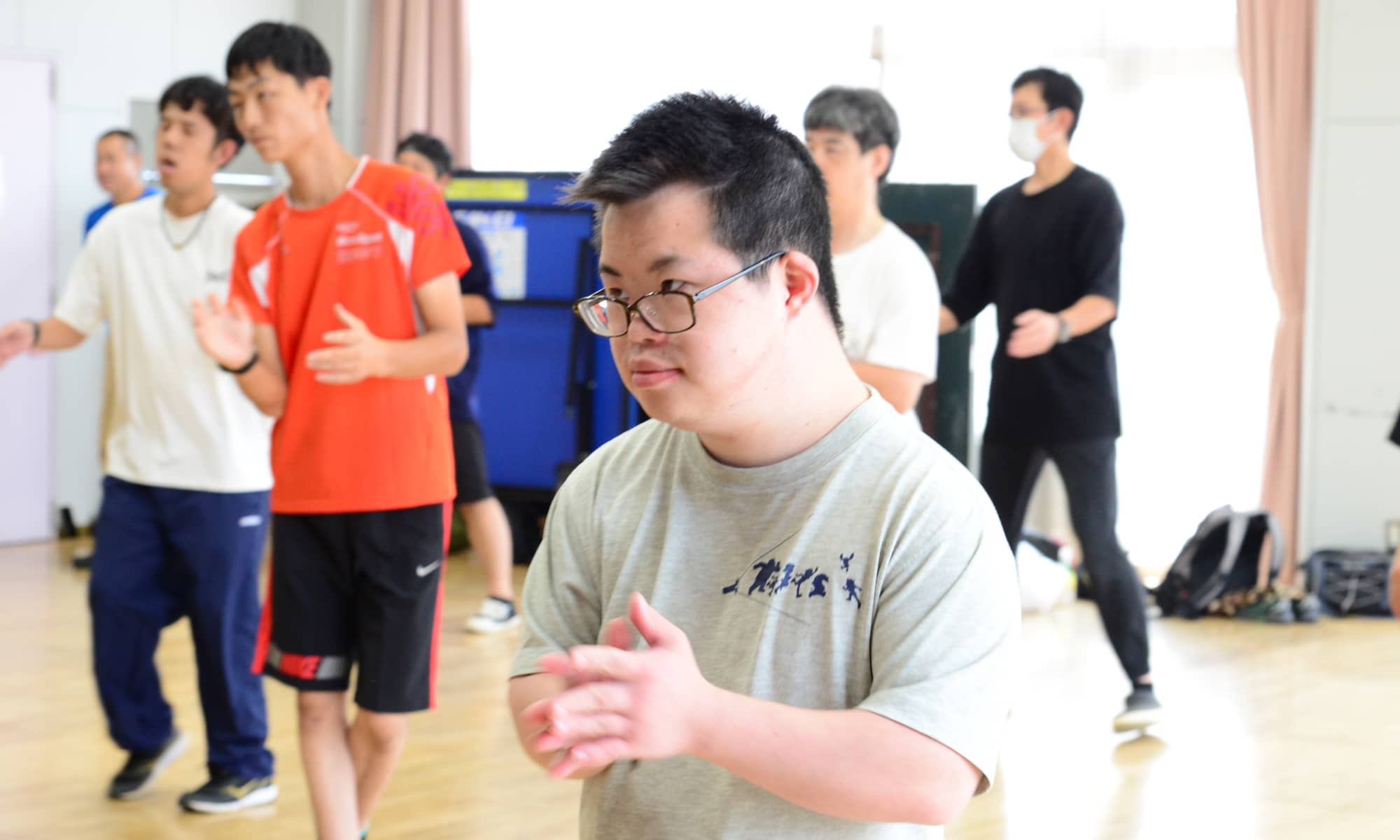 メガネをかけた男性が真剣な表情で手を動かしている。後ろにはダンスをする数名の人々が映っている