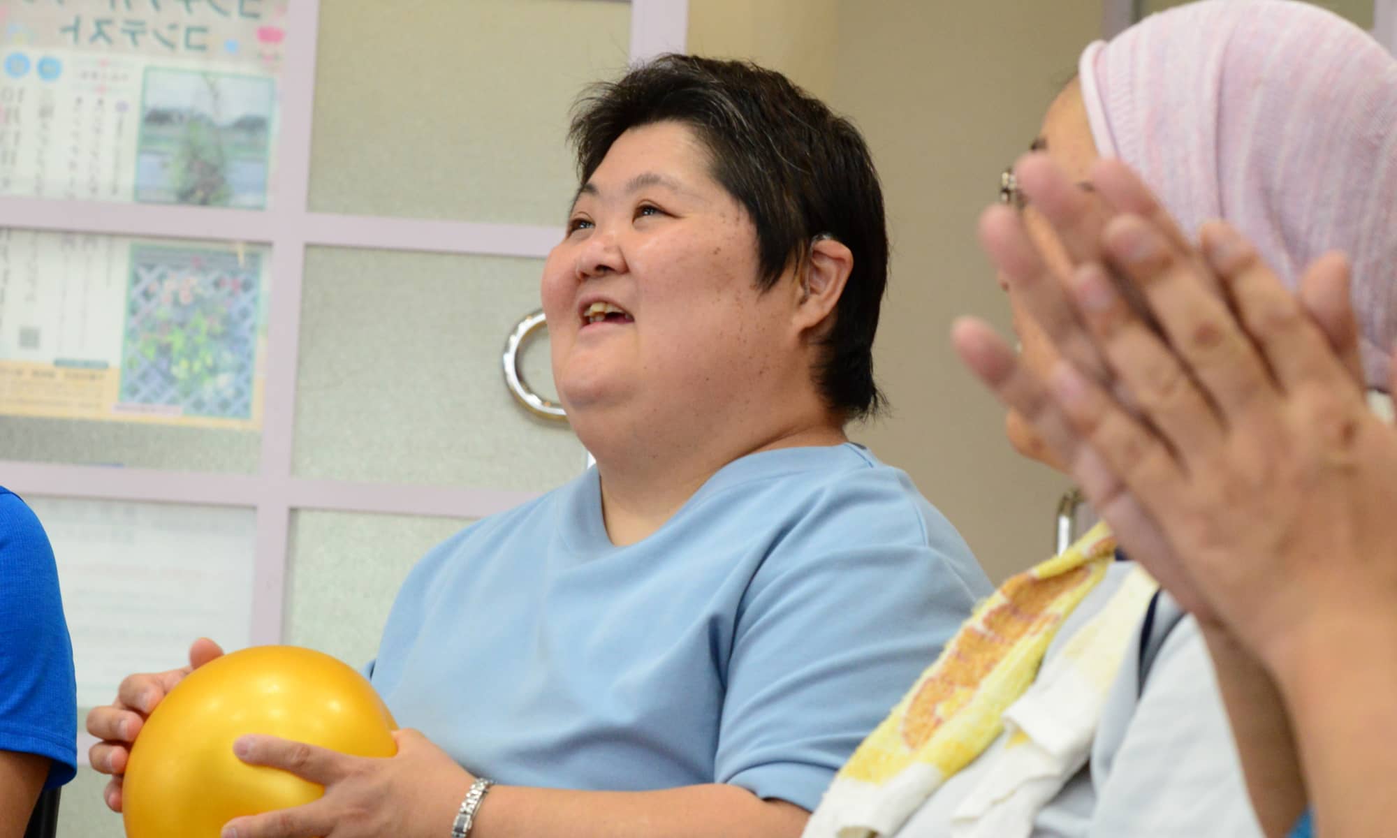 黄色いボールを持ちながら笑顔を見せる女性の様子。周囲の人々と楽しげに活動している場面