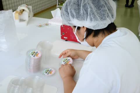 作業台で容器にシールを貼る帽子とマスク姿の作業員