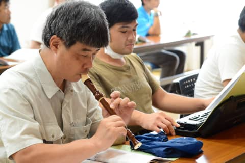 リコーダーを演奏する男性と隣でパソコンに向かう男性